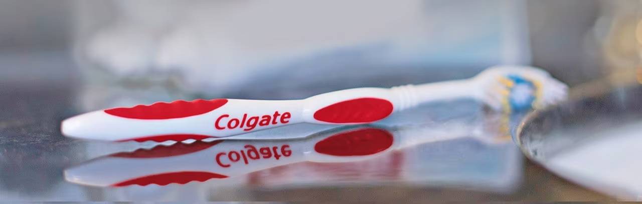 White and red Colgate manual toothbrush lying on a reflective surface, emphasizing daily oral hygiene to prevent cavities and tooth decay