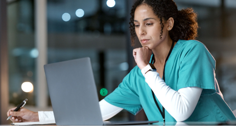 Female healthcare professional in teal scrubs working on a laptop and taking notes